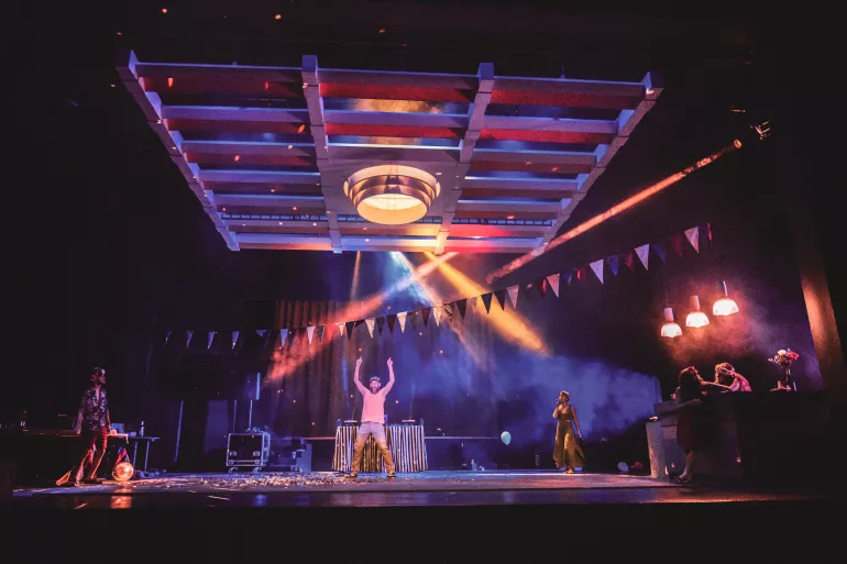 Une scène traversée par une guirlande de fanions et éclairée de lumières violettes et rouges est surplombée d’un quadrillage industriel. Au milieu de ce quadrillage, un grand spot jaune. En dessous de celui-ci, un homme lève les bras et le pointe du bout des doigts, il semble faire la fête. Des confettis sont au sol ainsi qu’un ballon de baudruche. A la gauche de cet homme, une femme avec une couronne de fleurs semble faire son entrée en chantant, micro devant la bouche. 