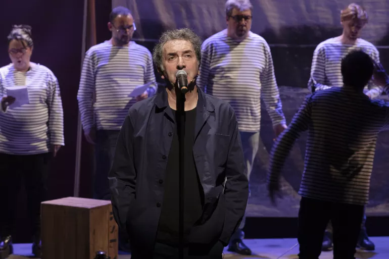 Un homme au premier plan semble chanter devant un micro. Il est habillé de gris foncé. Dans le fond en flou derrière lui on aperçoit cinq chanteurs tenant des papiers vêtus de marinières.  