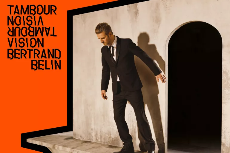 L’image est coupée à son premier tier gauche par une écriture noire déformé sur un fond orange, il est écrit ’Tambour vision Bertrand Belin’’. L’autre partie de l’image est occupée par un homme en costume penché légèrement vers l’avant. Le mur derrière lui présente une alcôve sombre. 