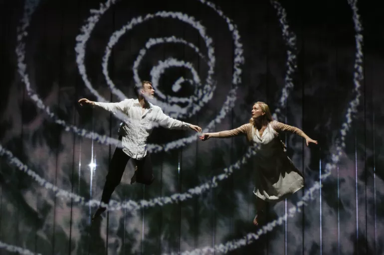 Un homme et une femme semblent danser ensemble. Ils ont tous les deux l’air surpris et l’homme regarde vers le haut. La femme fixe l’homme. Ils sont par endroits comme floutés car au premier rang une spirale blanche flotte sur eux. 