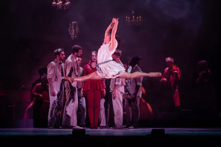 Un groupe d’homme en arrière-plan autour d’une femme habillée en rouge, elle a un micro et la bouche entrouverte. A leur gauche, un musicien dans le noir joue du violon. Au premier plan, une femme est en plein saut, grand écart. Elle porte une robe blanche et des pointes. Ses bras sont en l’air.