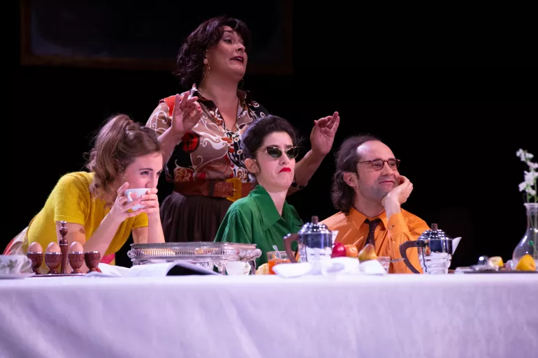 Trois personnes se tiennent assises au premier plan et une dame debout derrière. Elle est en pleine explication : bras soulevés et bouche entrouverte. Une des trois personnes devant est un homme, crâne dégarni et cheveux à l’arrière. Il a un air rêveur, des lunettes et affiche un sourire. La femme à côté de lui est excentrique, elle a des lunettes noires en yeux de chat, une bouche rouge et un chignon. Sa peau blanche contraste avec ses cheveux bruns et son chemisier vert.