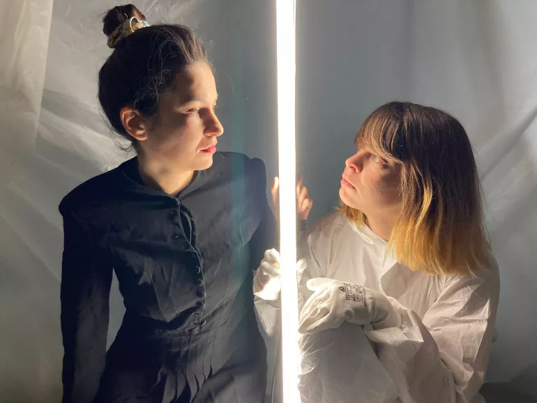 Séparées par un néon lumineux qu’elles tiennent dans leurs mains, deux femmes de profil se regardent yeux dans les yeux. À droite, une femme dont les cheveux blonds tombent sur ses épaules, est habillée d’une combinaison blanche et portent des gants de sécurité. À gauche, une femme plus grande dont on voit à peine les cheveux, porte une robe noire à boutons. 