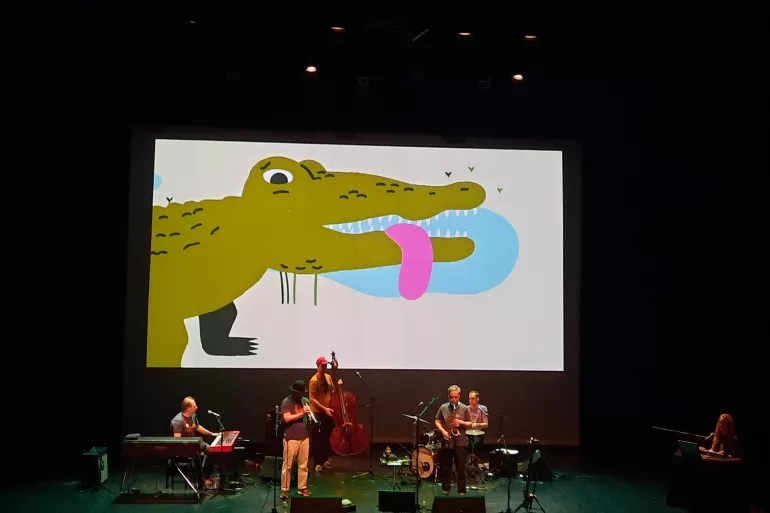 Sur une scène, se trouve un ensemble de musiciens habillés de tee-shirts et de pantalons dans les tons crème et gris. Au premier plan, se trouve un saxophoniste et un trompettiste. Au second plan, à gauche, un homme est au clavier puis au centre un contrebassiste, et à droite un batteur. Ils sont surplombés par un écran qui présente un dessin enfantin d’un crocodile tirant la langue. 