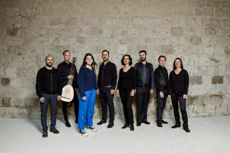 Huit femmes et hommes sont debout face à l’objectif, ils sont positionnés devant un mur ancien en pierres grises. Ils sont habillés en noir sauf une qui est en bleu clair. A sa droite, un homme tient une cithare en main. Ils regardent l’objectif avec un air neutre.  