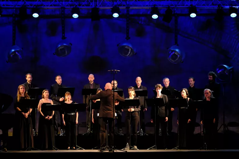Plusieurs chanteurs et chanteuses sont regroupés en deux rangs de deux niveaux. Derrière eux un fond bleu nuit teinté de noir semble se mouvoir. Ils sont habillés en costumes et en robes noires. Certains ont leurs bouches entrouvertes, ils chantent. Au premier plan, un homme est de dos les bras écartés c’est le chef de chœur.  