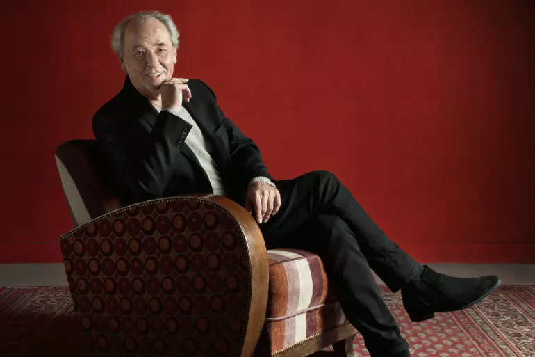 Un homme âgé, le chanteur Maxime Le Forestier, semble nous regarder et nous sourire. Il est habillé d’un costume noir et blanc et est assis sur un fauteuil ancien de cuir marron. Son buste est tourné vers l’objectif, il croise les jambes, sa main gauche est posée sur ses cuisses et sa main droite effleure son menton. Au sol, se trouve une moquette ornementée de couleurs rouges et vertes. Le mur derrière lui est rouge. 