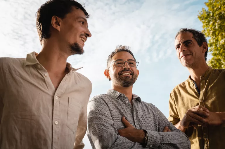 Trois hommes forment un demi cercle. Ils sont tout trois en chemise légères et pris en photo en contre bas. Au dessus d'eux le ciel est bleu et le soleil se reflète sur leurs visages. L'un deux, au centre, est plus petit et porte des lunettes rondes et une barbe, les deux autres sont plus grands.