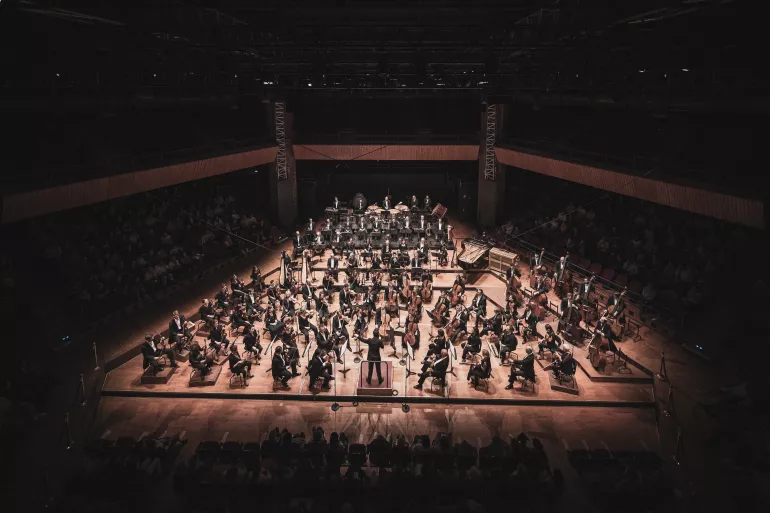 Dans une mise en lumière ciblée se trouve un orchestre assez grand sur une scène en bois de forme triangulaire. Au centre, on devine le chef d’orchestre, les bras levés. Autour de lui se trouvent les musiciens. On peut deviner dans l’ombre les spectateurs autour de la scène et aux balcons. 