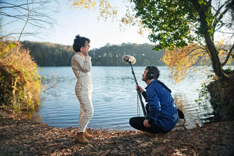 Une femme et un homme sont de profil devant un lac entouré de végétation. Ils posent leurs pieds sur des feuilles de chêne qui jonchent le sol. La femme debout est habillée d’une combinaison blanche et touche de sa main droite le casque audio qu’elle porte sur sa tête. L’homme accroupi face à elle la regarde. Il est lui aussi équipé d’un casque audio. Il est habillé d’une veste bleue et tient un micro-perche équipée d’une bonnette en fourrure dans ses mains. 