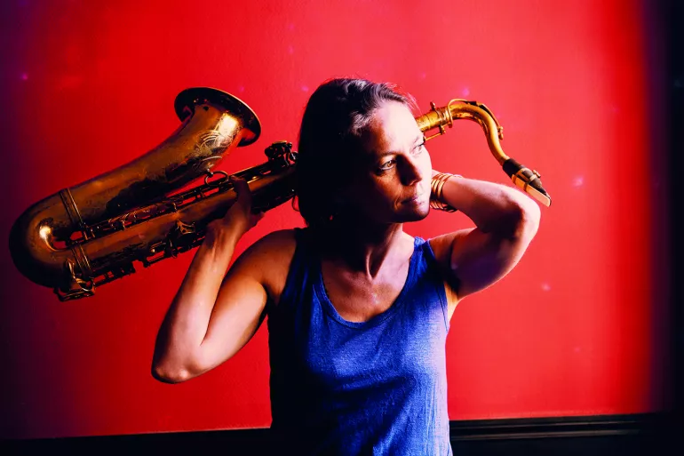 Sur un fond rouge, une femme dans un plan américain se tient face à nous, la tête tournée vers la droite. Habillée d’un jean et d’un débardeur bleu, elle tient derrière sa tête, à l’aide de ses deux mains, un saxophone. 