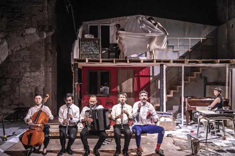 Au centre d’un décor d’une maison délabrée se trouve une femme allongée semblant mimer la mort. Elle est habillée dans des couleurs vives, le rose et le bleu de ses vêtements contrastent avec l’ensemble du décor. Debout au-dessus d’elle, un homme en costume la pointe du doigt. De part et d’autre de la scène, deux musiciens sont tournés vers le corps de la femme, ils portent chacun un instrument et sont tous deux habillés d’un haut blanc et d’un pantalon noir.    