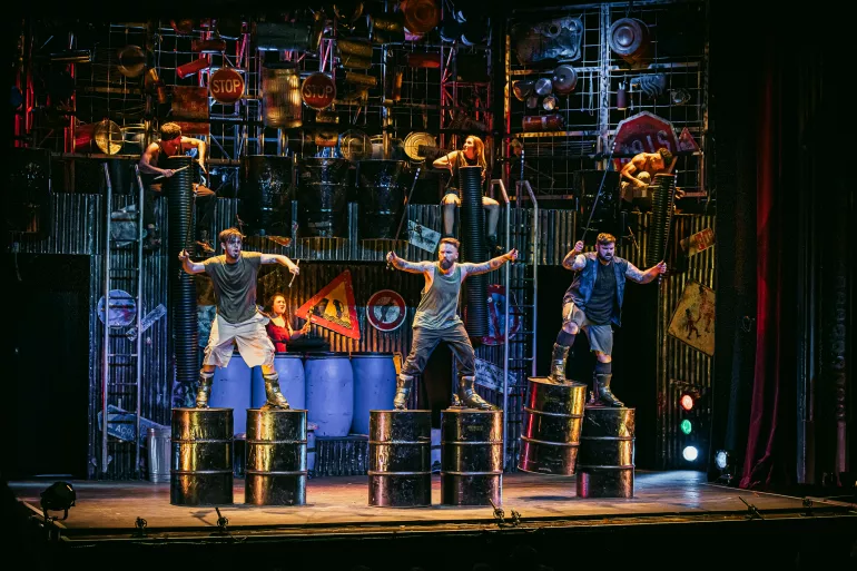 Sept percussionnistes sont répartis sur trois niveaux dans un décor industriel sombre. Des panneaux de signalisation de chantier sont dispersés sur la scène. Au premier plan, trois hommes sont debout, ils ont des barils en fer accrochés à leurs pieds. Ils ont les bras en l’air et bougent aux rythmes de leurs percussions. En arrière-plan, une femme joue des percussions sur des barils en plastique. 