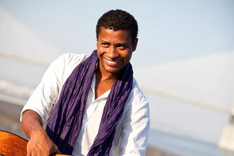 Un homme de couleur sourit de toutes ses dents avec le regard en biais. Il porte une écharpe violette et une chemise blanche. Il a également un collier en argent. 