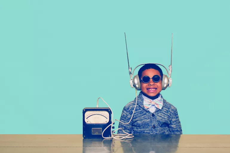 Un enfant de couleur au centre de l’image sourit de toutes ces dents. Il a sur les oreilles un casque audio surmonté de deux antennes. Le casque est relié à un poste de type ancien. L’enfant porte un nœud papillon et des lunettes. Le fond est bleu turquoise. 