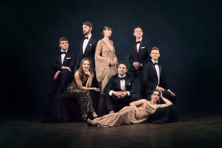 Un groupe d’hommes et de femmes posent dans une composition pyramidale. Respectivement cinq hommes, habillés de costumes noirs avec un nœud papillon et trois femmes vêtues de robes beiges et scintillantes. Ils posent tous dans une position différente. Trois d’entre eux sont debout, quatre sont assis sur des supports recouverts de tissus brillants s’apparentant à du satin et une femme, à leurs pieds, est allongée sur son flanc gauche. Seulement deux d’entre eux regardent l’objectif. 