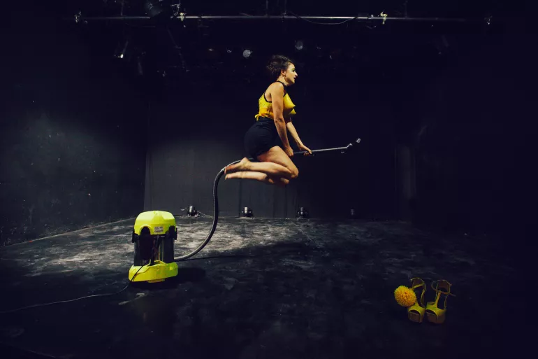 Sur scène, une femme habillée d’un short noir et d’un corset jaune chevauche le manche d’un aspirateur telle une sorcière. Sa main droite tient le manche et elle place sa main gauche sur son front, en guise de visière. Elle regarde au loin comme si elle cherchait quelque chose. 