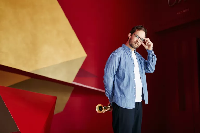 À droite d’un fond déstructuré géométrique de couleur jaune et rouge se trouve un homme qui nous regarde. Il est barbu, habillé d’une chemise bleue ouverte sur un t-shirt blanc et d’un pantalon bleu foncé. Sa tête est légèrement inclinée, avec sa main gauche, il touche les branches de ses lunettes comme s’il s’apprêtait à nous saluer. Avec sa main droite il tient derrière son dos son saxophone. 