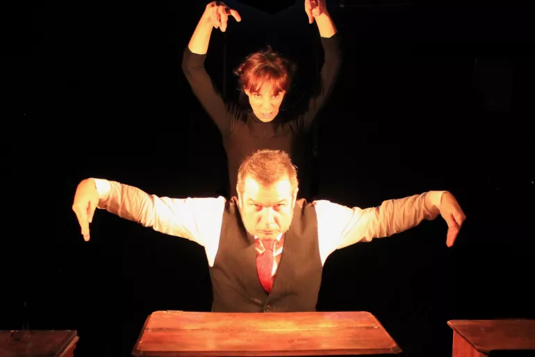 Un homme au premier plan un peu fort. Il porte un gilet, chemise blanche et cravate rouge. Ses yeux nous fixent, il est légèrement penché vers l’avant, ses bras levés comme un oiseau prêt à s’envoler.